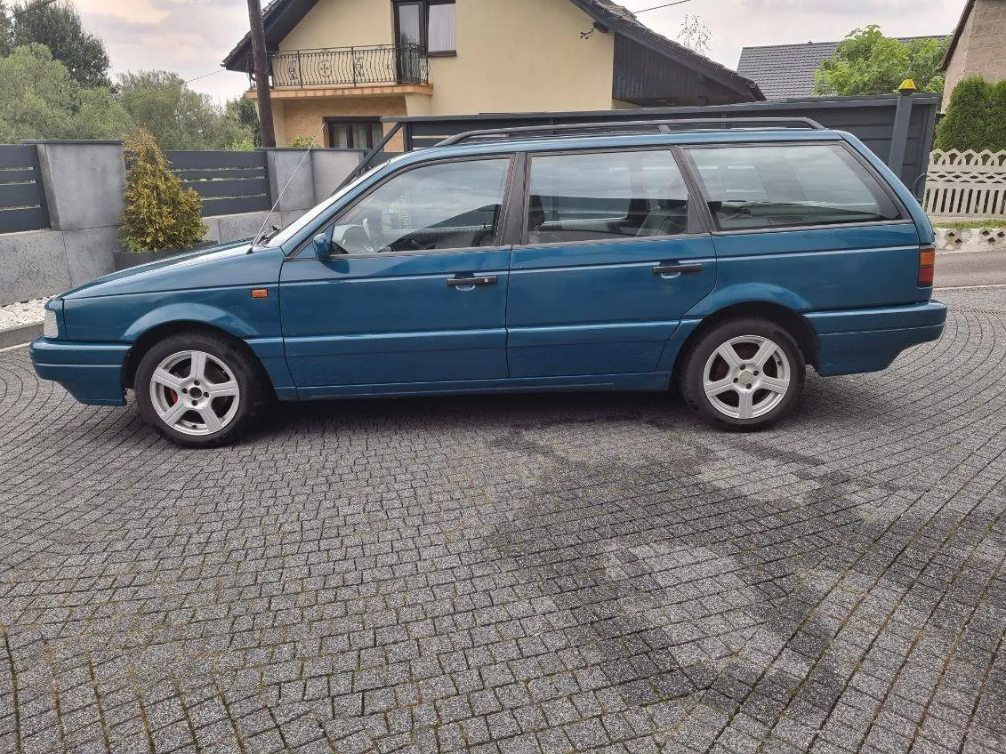 Niebieski samochód typu kombi zaparkowany na brukowanej nawierzchni przed domem o jasnym kolorze z balkonem i czarnym ogrodzeniem. W tle widoczne drzewa, krzewy oraz białe ogrodzenie. Samochód posiada dachówkę i srebrne felgi. Niebo jest pochmurne.