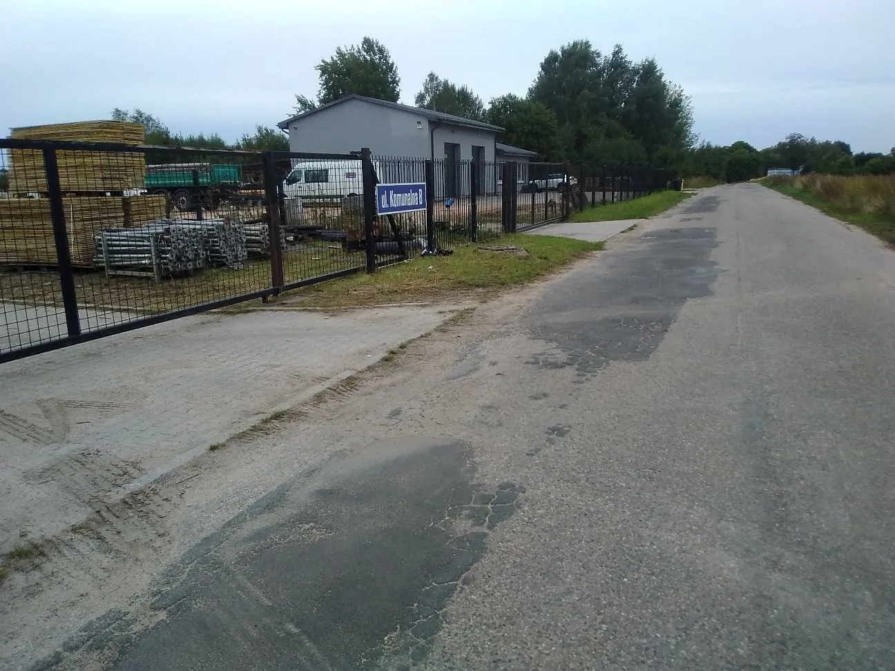 Widok na asfaltową drogę prowadzącą w głąb obrazu. Po lewej stronie znajduje się ogrodzony teren z czarnego metalowego płotu, za którym widoczne są stosy żółtych materiałów budowlanych, metalowe konstrukcje oraz szary, parterowy budynek gospodarczy. Na bramie wjazdowej zawieszona jest niebieska tabliczka z białym napisem: 'ul. Komunalna 8'. W tle widać zielone drzewa pod jasnym, zachmurzonym niebem.