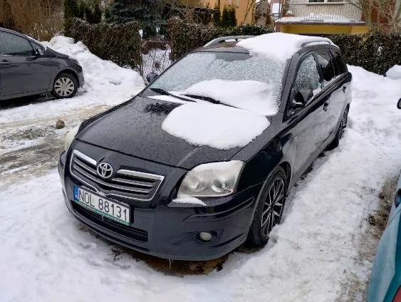 Czarny samochód Toyota kombi pokryty śniegiem, zaparkowany na śnieżnej nawierzchni. Widoczna tablica rejestracyjna NOL 88131. W tle inne pojazdy, krzewy i domy. Śnieg leży na dachu i maskie pojazdu, a także na ziemi.