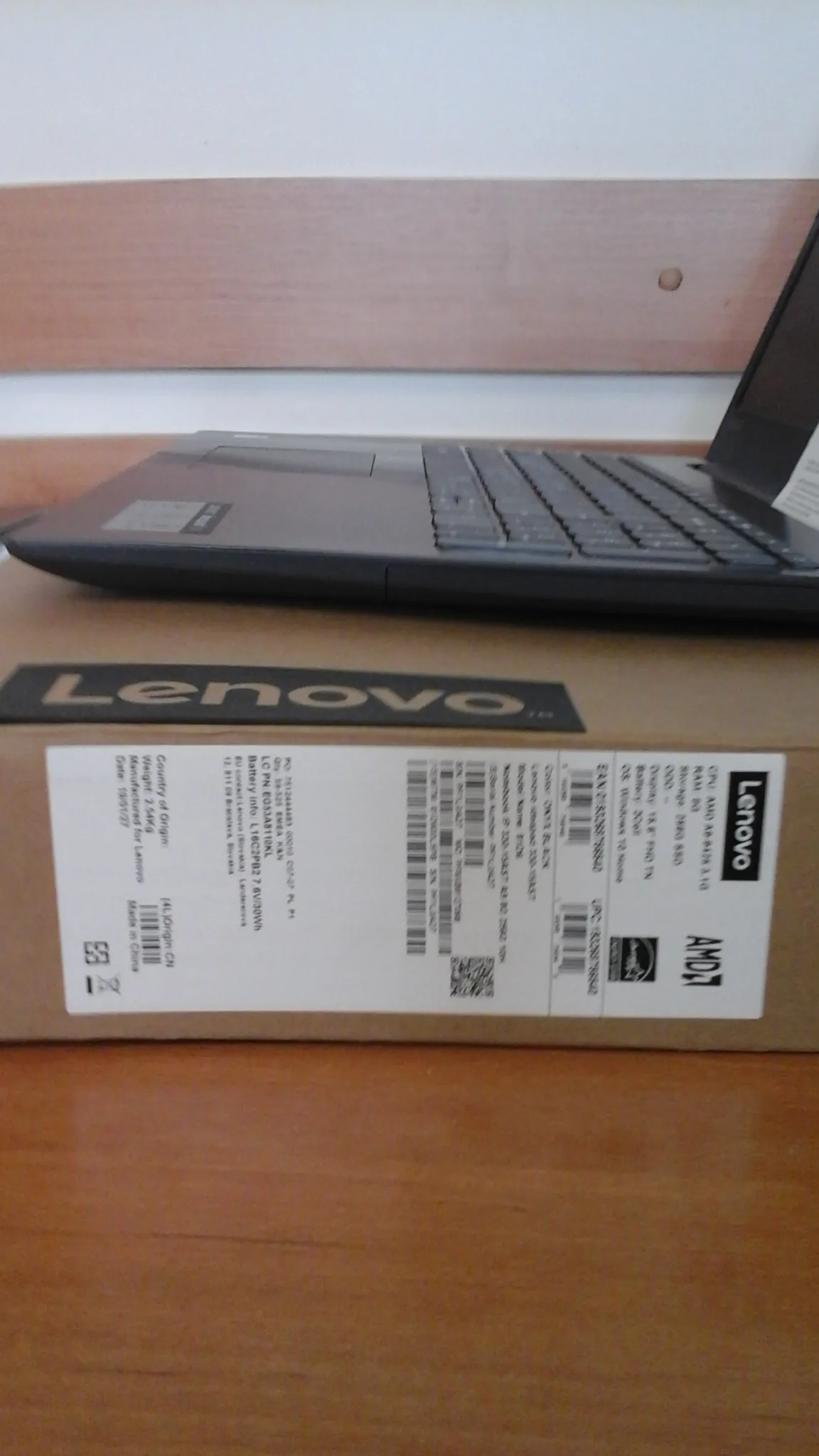 Zbliżenie na kartonowe opakowanie laptopa Lenovo z etykietą zawierającą specyfikację techniczną. Na etykiecie widoczne są napisy: 'Lenovo', 'AMD', dane dotyczące procesora (CPU: AMD A4-9125 2.30), pamięci RAM (4GB), pamięci masowej (256GB SSD), ekranu (14.0" FHD TN), baterii (2Cell), systemu operacyjnego (Windows 10 Home), kody kreskowe, informacje o pochodzeniu (Country of Origin: CN, Made in China) oraz waga (2.54kg). Po lewej stronie widoczna jest część klawiatury laptopa i drewniana powierzchnia.