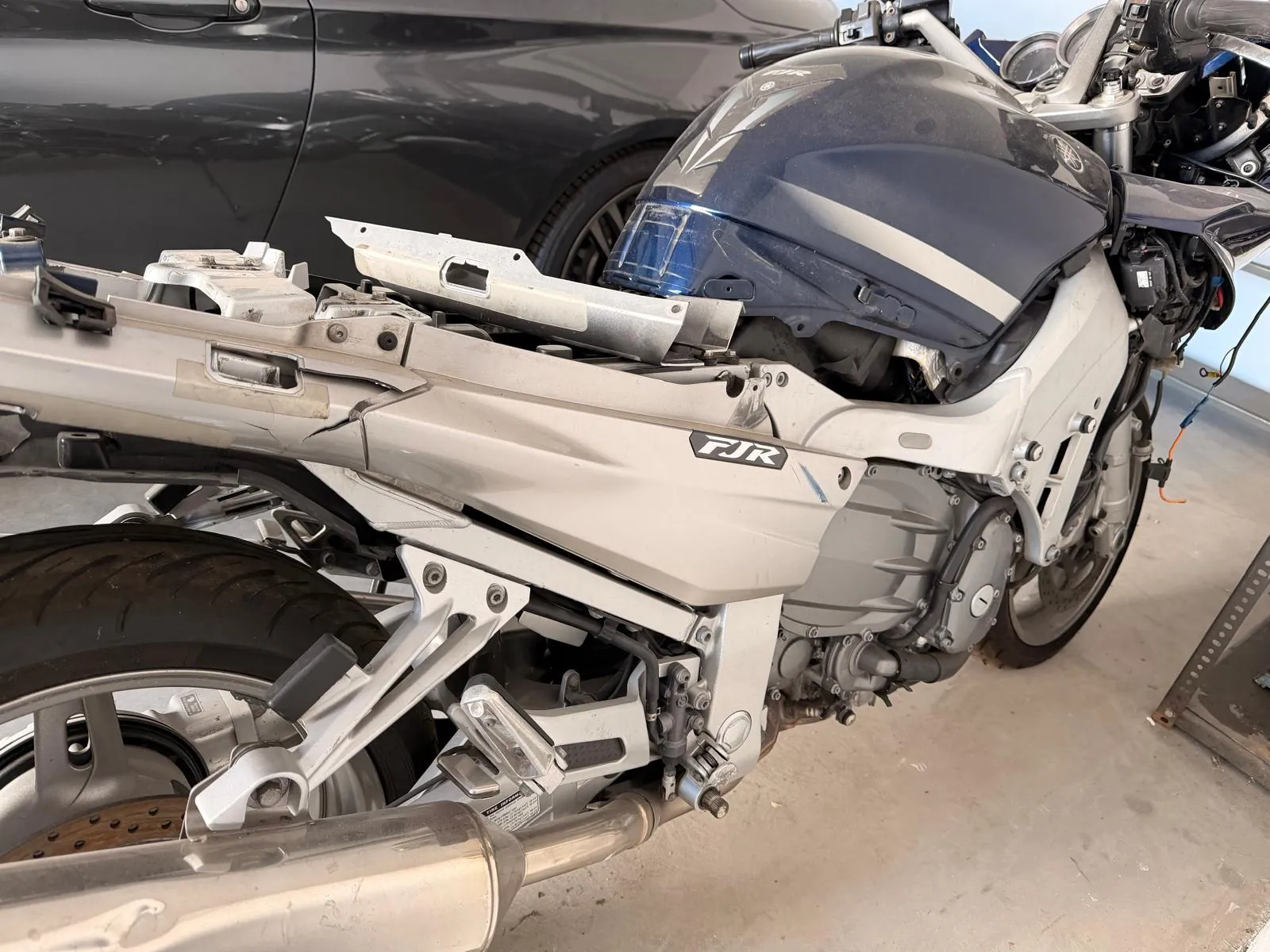 Zbliżenie na prawy bok srebrno-niebieskiego motocykla marki Yamaha z widocznym napisem FJR na owiewce. Motocykl jest częściowo rozebrany, brakuje niektórych elementów plastikowych osłon. W tle widoczny jest fragment ciemnego samochodu.