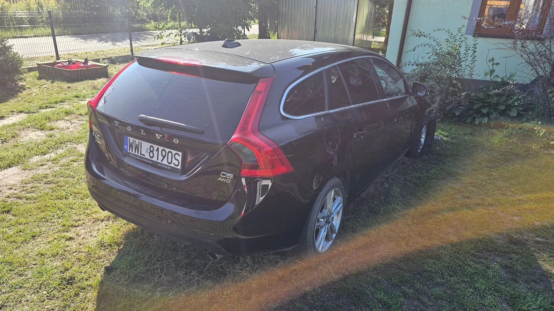 Czarny samochód marki Volvo w nadwoziu kombi, zaparkowany na trawiastym podjeździe. Widoczny jest tył i prawy bok pojazdu. Tablica rejestracyjna ma numer WWL 8190S. Na klapie bagażnika widnieją napisy 'VOLVO' oraz oznaczenie modelu 'D5 AWD'. W tle widać zieloną roślinność, ogrodzenie oraz fragment budynku.
