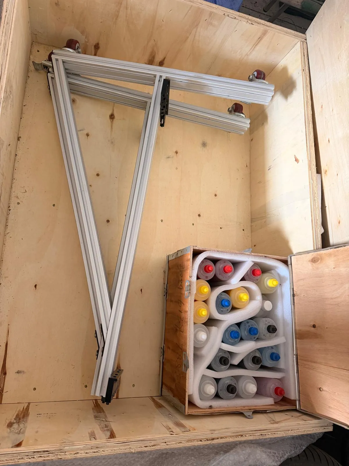 Wnętrze drewnianej skrzyni transportowej z zapakowanymi elementami. W górnej części znajduje się biała, metalowa konstrukcja z profili aluminiowych w kształcie litery V z poziomą belką. Na dole skrzyni znajduje się mniejszy drewniany pojemnik z białym styropianem, w którym umieszczone są liczne plastikowe butelki z kolorowymi cieczami (żółtymi, czerwonymi, niebieskimi i białymi).