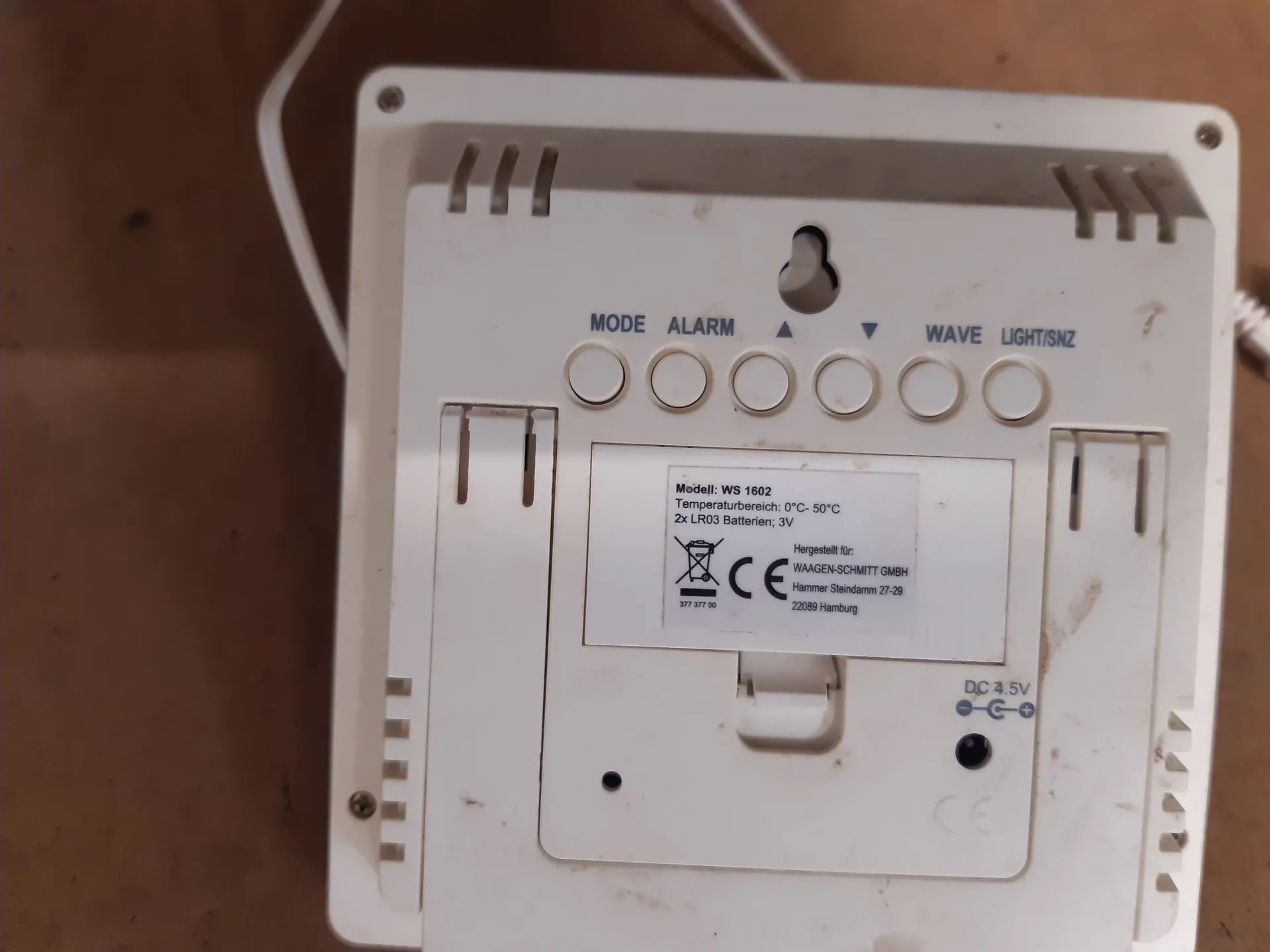 Tylna obudowa białego urządzenia elektronicznego z rzędem sześciu okrągłych przycisków. Nad przyciskami znajdują się napisy: MODE, ALARM, strzałka w górę, strzałka w dół, WAVE oraz LIGHT/SNZ. Poniżej znajduje się biała etykieta z tekstem: 'Modell: WS 1602, Temperaturbereich: 0°C- 50°C, 2x LR03 Batterien; 3V'. Na etykiecie widnieją również symbole przekreślonego kosza na śmieci, znak CE oraz dane producenta: 'Hergestellt für: WAAGEN-SCHMITT GMBH, Hammer Steindamm 27-29, 22089 Hamburg'. W prawym dolnym rogu znajduje się gniazdo zasilania z oznaczeniem 'DC 4.5V'.