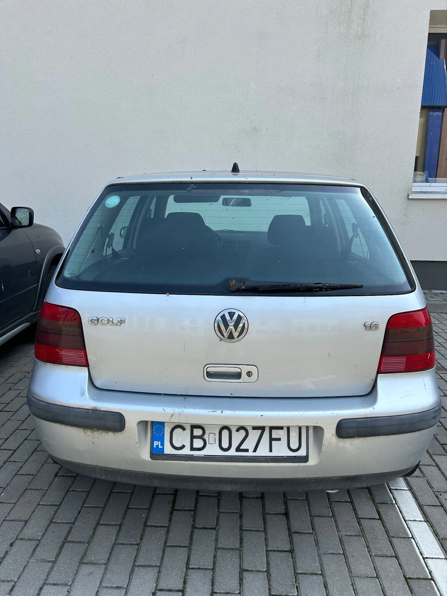 Tylny widok srebrnego samochodu marki Volkswagen Golf. Na klapie bagażnika widoczne jest logo Volkswagena oraz napis GOLF i oznaczenie 1.6. Samochód posiada polską tablicę rejestracyjną o numerze CB 027FU. Auto zaparkowane jest na kostce brukowej przed jasną ścianą budynku.