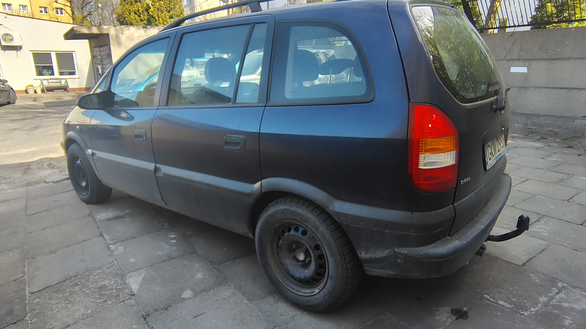 Ciemnoniebieski samochód typu minivan marki Opel, brudny, z dachowym bagażnikiem i holownikiem. Widoczna tablica rejestracyjna z napisem GKM 29. Samochód zaparkowany na brukowanej powierzchni. W tle budynek z oknami, klimatyzatorem, drzewami i betonowym murem.
