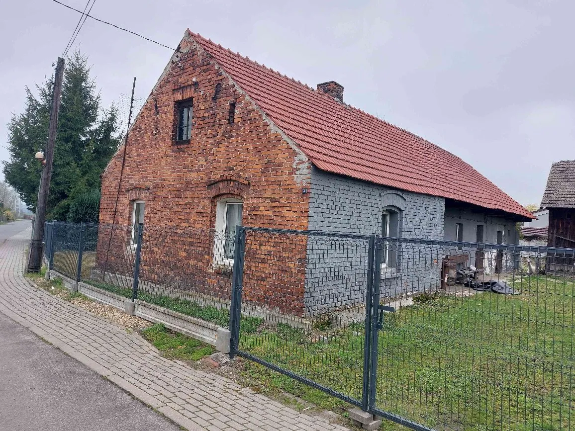 Jednokondygnacyjny budynek mieszkalny z czerwonej cegły z dwuspadowym dachem pokrytym czerwoną dachówką. Część budynku po prawej stronie jest pomalowana na szaro. Dom znajduje się za ciemnozielonym metalowym ogrodzeniem z siatki. Przed domem przebiega chodnik z kostki brukowej oraz asfaltowa droga. W tle widoczne są drzewa i pochmurne niebo.