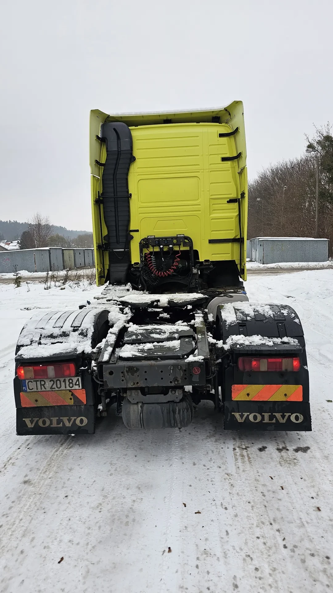 Zdjęcie przedstawia ciężarówkę Volvo z otwartą żółtą kabiną, pokrytą śniegiem. Widoczne są czarne elementy, tablica rejestracyjna CTR 20184, pomarańczowo-żółte odblaski oraz śnieg na częściach pojazdu. Tło: śnieżna droga, drzewa i budynki.