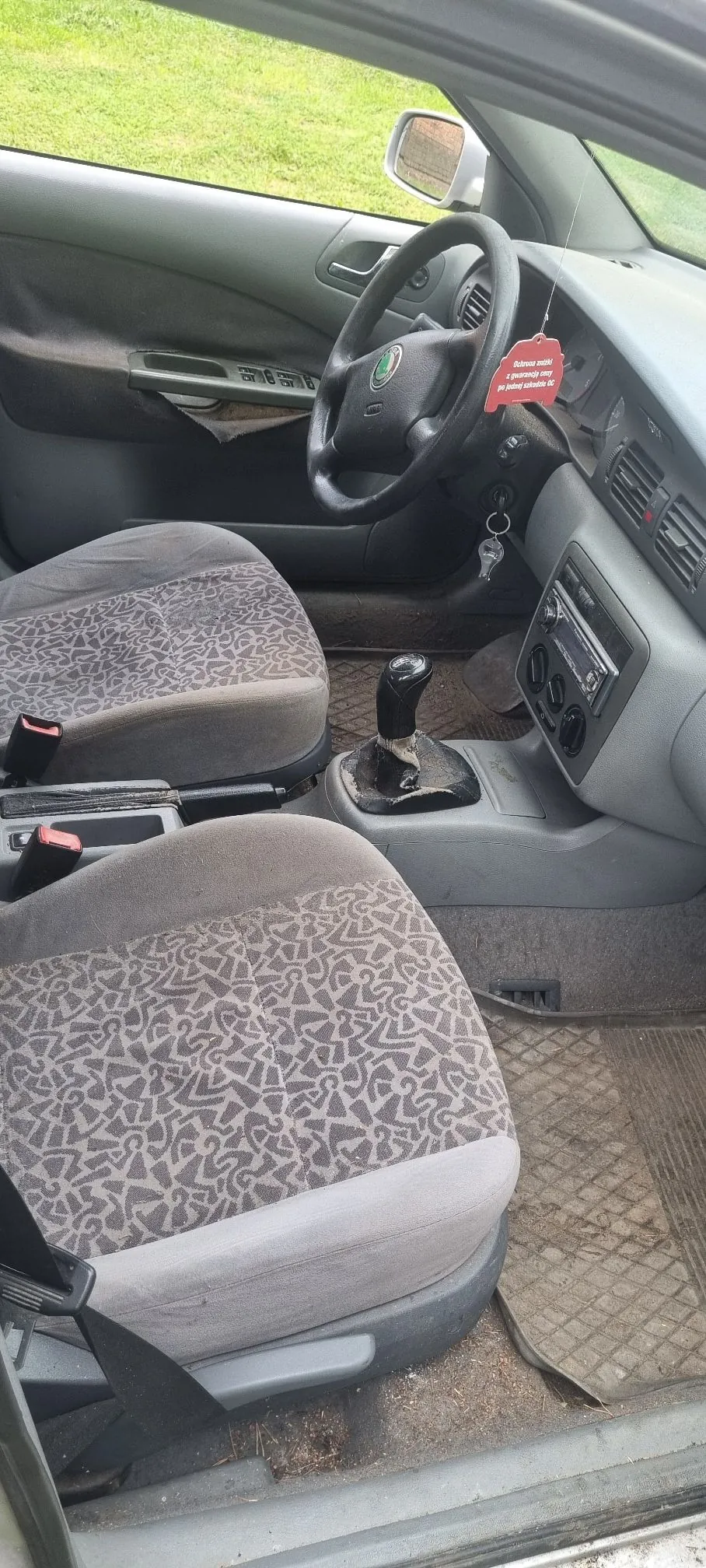 Wnętrze samochodu widziane z perspektywy otwartych drzwi pasażera. Widoczne są dwa przednie fotele z szarym, wzorzystym obiciem, kierownica z logo marki Skoda, drążek zmiany biegów oraz deska rozdzielcza z radiem i nawiewami. Na lusterku wstecznym wisi czerwona zawieszka z białym napisem. Podłoga wyłożona jest gumowymi dywanikami.
