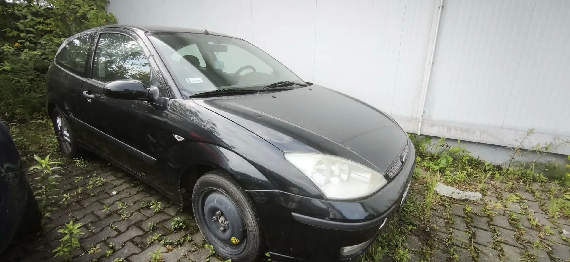 Czarny samochód osobowy typu hatchback zaparkowany na kostce brukowej porośniętej trawą, na tle białego ogrodzenia z paneli. Pojazd jest widoczny z przodu i z boku, z lekkim kątem z góry.