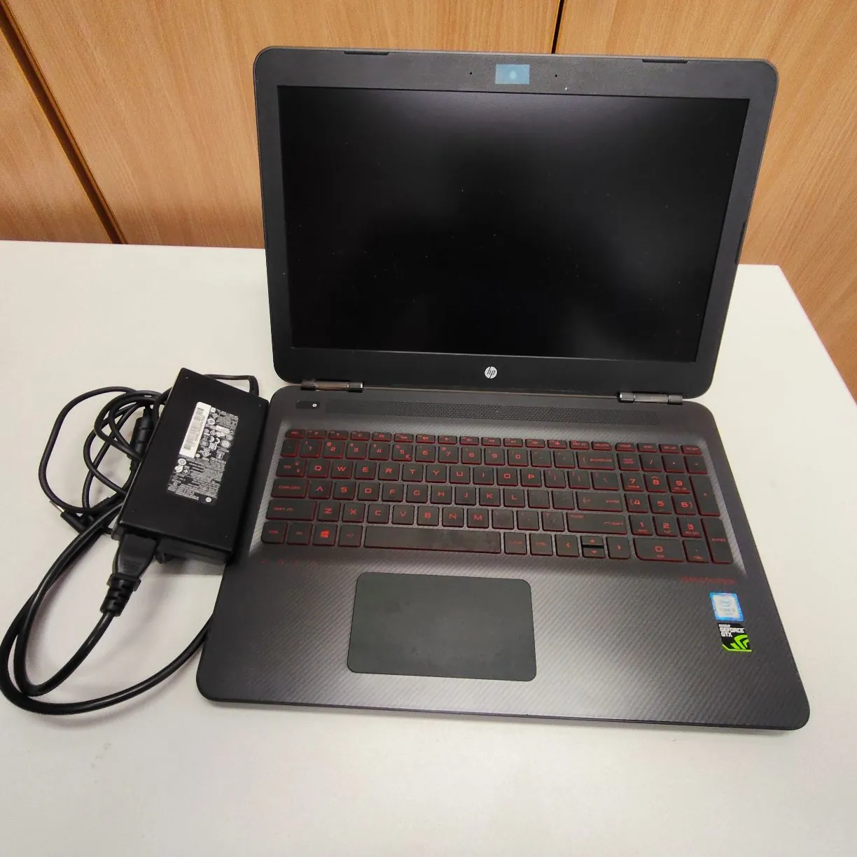 Otwarty laptop marki HP w kolorze czarnym z czerwoną podświetlaną klawiaturą, ustawiony na białym blacie. Obok laptopa, po lewej stronie, znajduje się czarny zasilacz z podłączonym kablem. Na obudowie laptopa, w prawym dolnym rogu, widoczne są naklejki producentów: Intel Core i7 oraz NVIDIA GeForce GTX.