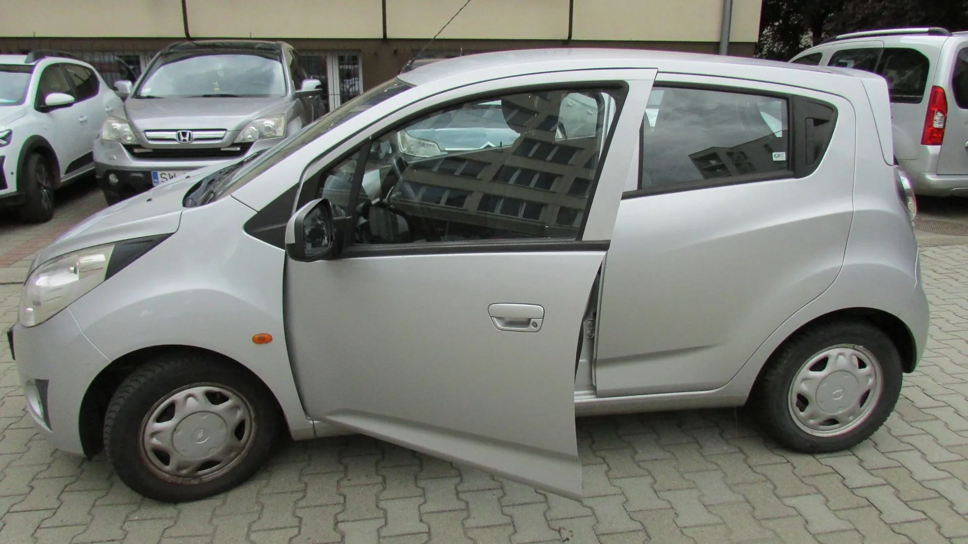 Srebrny samochód osobowy typu hatchback zaparkowany na kostce brukowej. Przednie lewe drzwi samochodu są otwarte. W tle widoczne są inne zaparkowane samochody: biały po lewej stronie oraz srebrny samochód marki Honda bezpośrednio za głównym pojazdem.