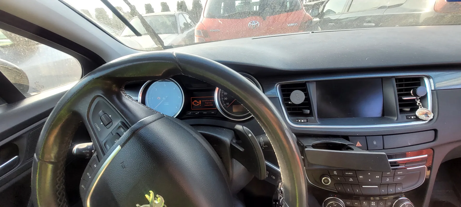 Wnętrze samochodu widziane z perspektywy kierowcy. Widoczna czarna kierownica z logo Peugeot, zegary przyrządów (w tym prędkościomierz i tachometr), cyfrowy wyświetlacz z pomarańczowym komunikatem ostrzegawczym. W centralnej konsoli ekran, klimatyzacja, przyciski sterujące i klucz wiszący na klipsie. Za przednią szybą zaparkowane samochody, w tym czerwony Toyota Yaris z widocznym logo i napisem "YARIS". Tekst na wyświetlaczu: "Uwaga: system nie działa prawidłowo".