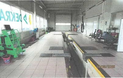 Wielka hala warsztatowa z podłogą z kafelków, zielonym urządzeniem po lewej stronie, widocznym napisem "DEKRA" na oknie, rowami w podłodze, krzesłami i narzędziami po prawej stronie oraz bramą garażową w tle. W prawym dolnym rogu data "24/08/2006".