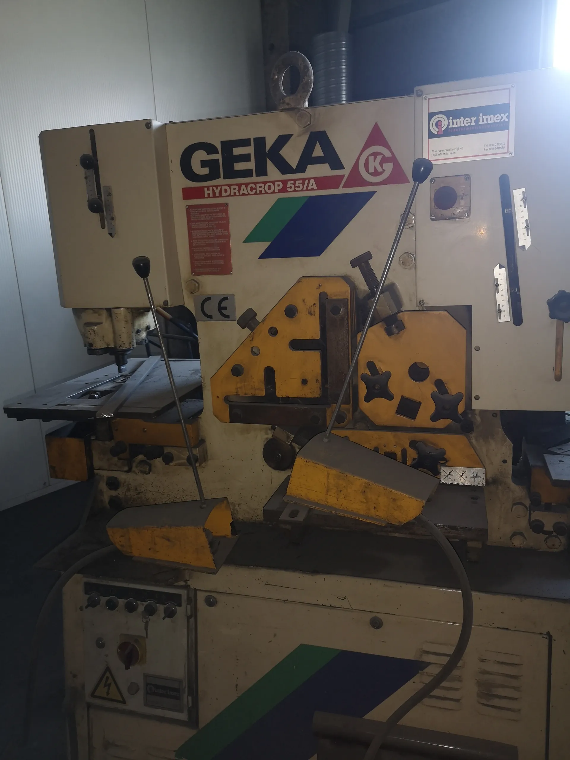 Przemysłowa maszyna GEKA HYDRACROP 55/A z widocznym logo KC w czerwonym trójkącie oraz zielono-niebieskim pasem. Na maszynie znajdują się żółte elementy mechaniczne, dźwignie z czarnymi uchwytami, czerwona tabliczka ostrzegawcza, znak CE oraz tabliczka z napisem 'inter imex'. W dolnej części widoczny jest panel sterowania z przyciskami i żółtym znakiem ostrzegawczym przed prądem. Maszyna umieszczona w pomieszczeniu przemysłowym z białymi ścianami i metalowymi elementami w tle.
