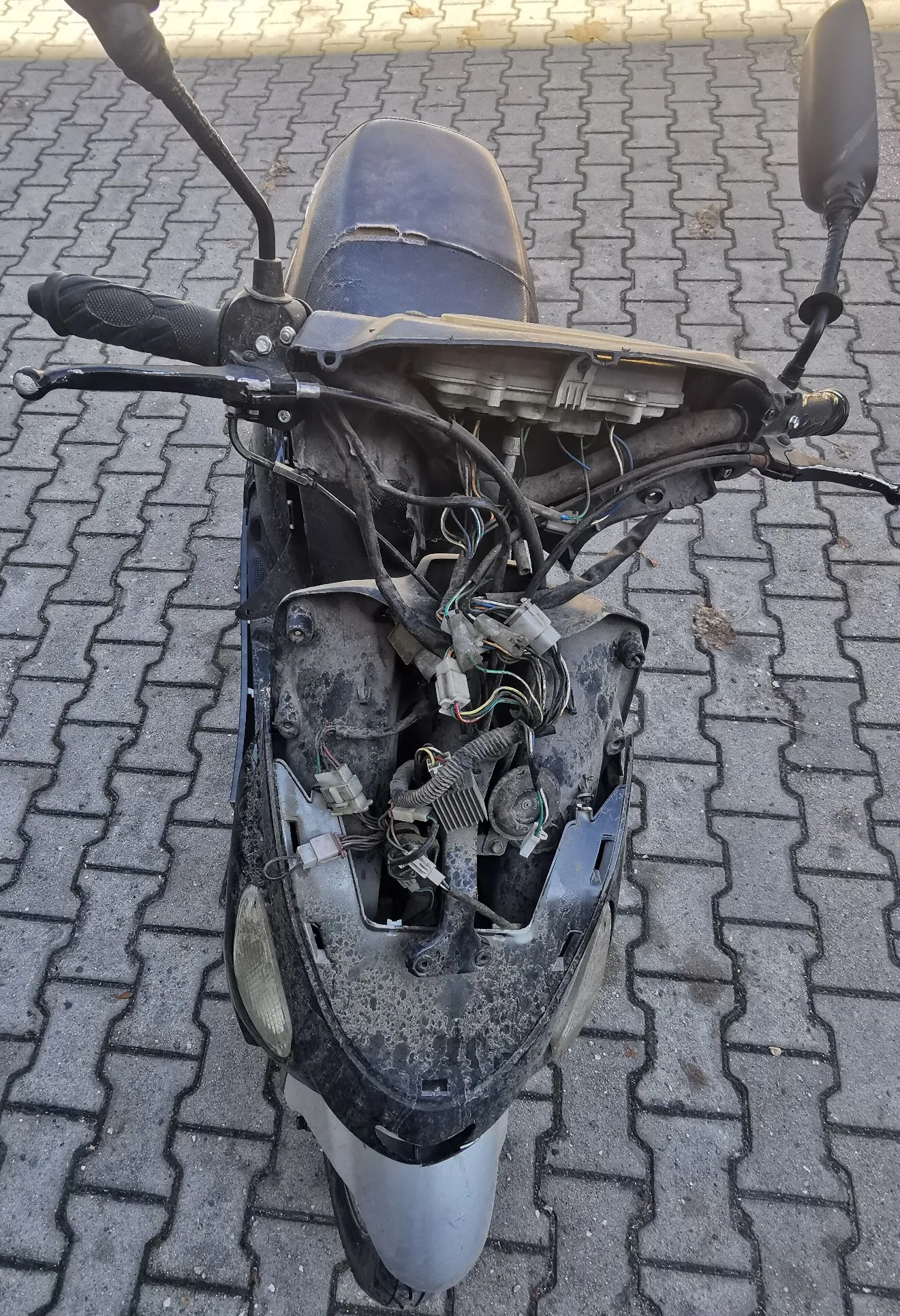 Widok z góry na ciemny skuter zaparkowany na kostce brukowej. Przednia obudowa skutera jest zdjęta, odsłaniając wewnętrzne komponenty, w tym wiązki przewodów elektrycznych, złącza i elementy ramy. Skuter posiada czarne lusterka i manetki.