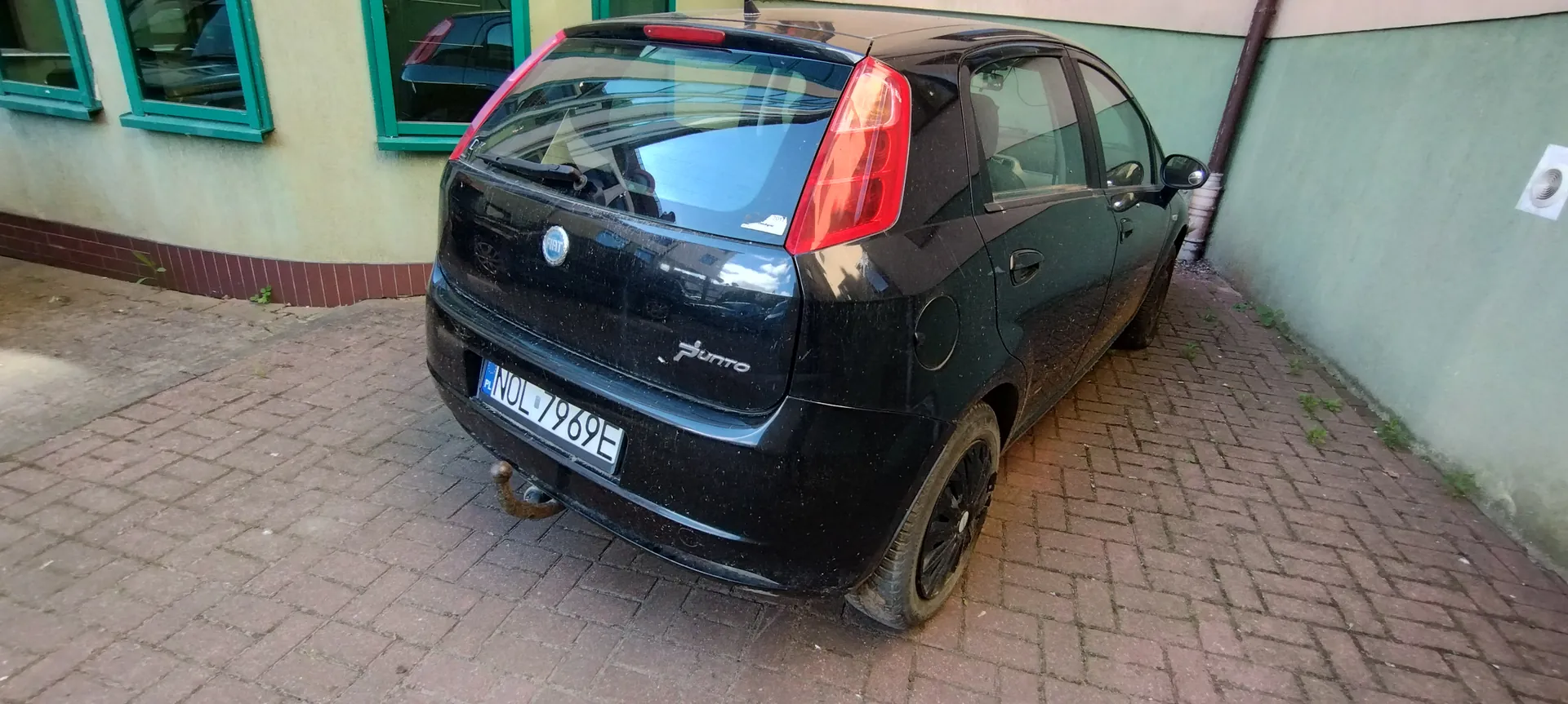 Czarny samochód Fiat Grande Punto zaparkowany tyłem do obserwatora na brukowanym podwórku. Widoczna tablica rejestracyjna PL NOL 7969E, czerwone światła tylnego, logo Fiata oraz napis "Punto" na bagażniku. Po lewej stronie budynku z zielonymi ramami okiennymi i beżowo-zieloną elewacją, po prawej zielona ściana z rurą odprowadzającą. Podłoga wyłożona czerwoną cegłą, na której rosną pojedyncze zioła.