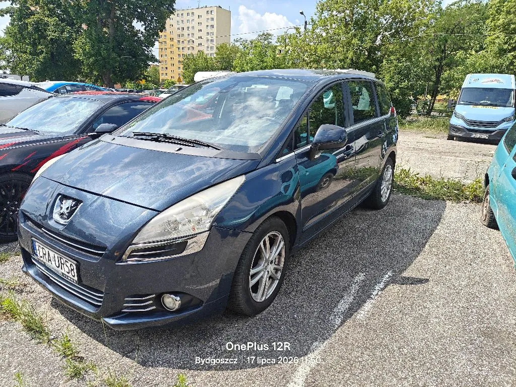 Ciemnoniebieski samochód marki Peugeot zaparkowany na asfaltowym placu. Pojazd jest widoczny z przodu i z boku. Na tablicy rejestracyjnej widnieje napis ERA UR58. W tle widać inne samochody, zielone drzewa oraz wysoki, żółty blok mieszkalny. Na dole obrazu znajduje się biały napis: OnePlus 12R Bydgoszcz 17 lipca 2024 11:50