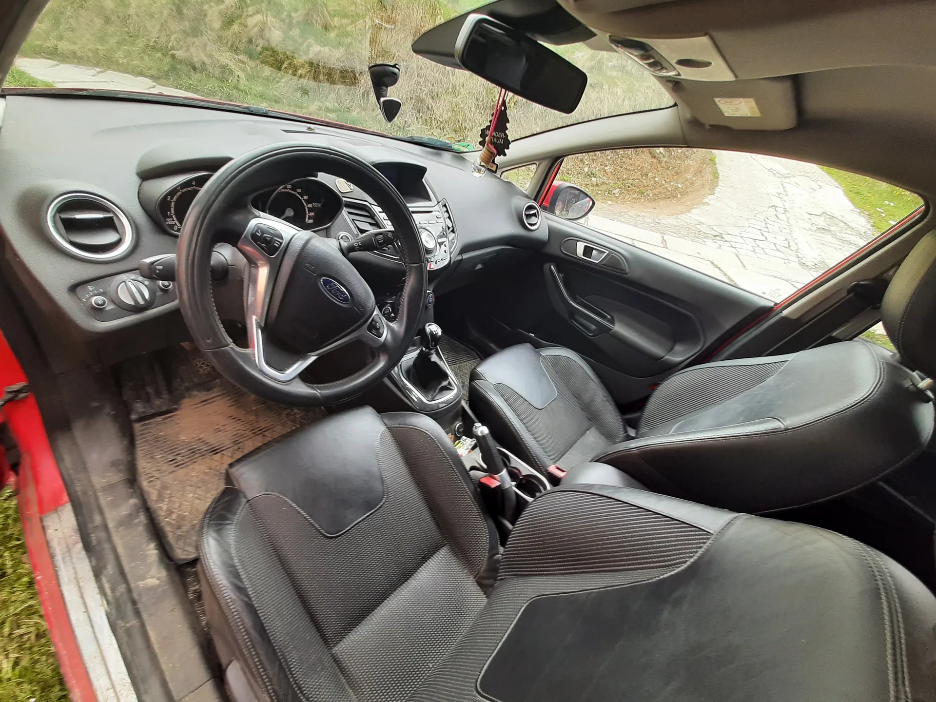 Wnętrze samochodu marki Ford z czarnymi siedzeniami o teksturze siatki, kierownicą z logo Ford, czarną deską rozdzielczą z licznikami i przyciskami. Drzwi kierowcy są otwarte, widoczna czerwona karoseria. W tle przez przednią szybę i otwarte drzwi widać trawę oraz brukowaną drogę. Lusterko wsteczne i wiszący na nim przedmiot są widoczne na suficie pojazdu.