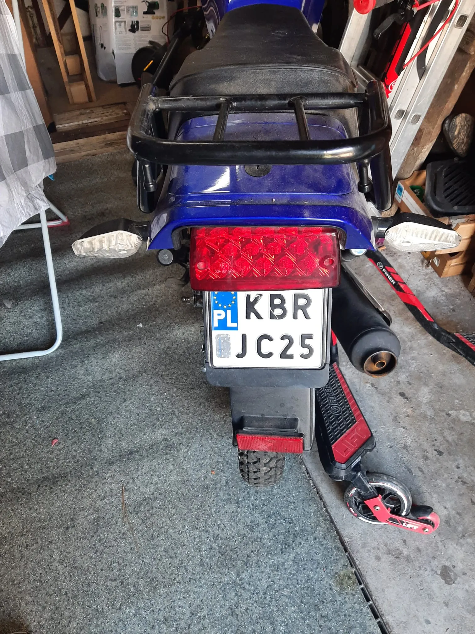 Zdjęcie przedstawia tył niebieskiego motocykla z czarnym siedzeniem i czerwonym światłem tylnym. Na motocyklu umocowana jest biała tablica rejestracyjna z niebieskim pasem po lewej stronie zawierającym napis "PL" oraz czarnymi literami i cyframi "KBR JC25". Po prawej stronie motocykla widoczny jest czerwono-czarny hulajnoga. Tło stanowi garaż z betonową podłogą, na której leżą różne przedmioty, w tym drabina, pudełka i części rowerowe. Po lewej stronie znajduje się szaro-biała tkanina na stojaku.