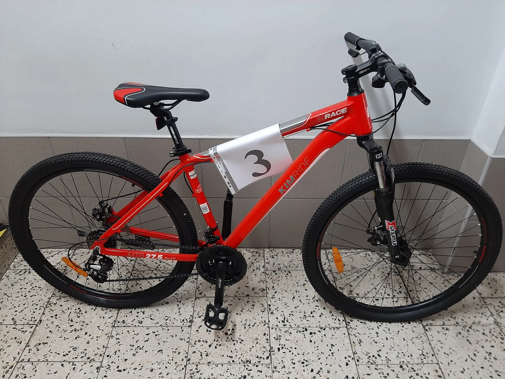 Czerwony rower górski marki KIMRIDE z napisem M RACE na ramie. Na ramie umocowany biały numer startowy z czarną cyfrą 3. Rower ma czarne opony, czarno-czerwone siedzisko oraz czarne kierownicę z hamulcami. Na dolnej części ramy widoczny napis MTB 27.5 ALLOY. Rower stoi na podłodze z białymi płytkami w czarne kropki, przed ścianą z szarymi płytkami na dole i białą powierzchnią na górze.