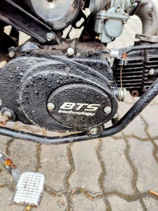 Zbliżenie na czarny silnik motocykla pokryty kroplami wody. Na okrągłej pokrywie widoczny biały napis BTS technology. W tle fragment chodnika z kamiennymi płytami oraz metalowa podnóżka z siateczkowym wzorem. Widoczne są również elementy konstrukcyjne silnika, takie jak przewody i śruby.