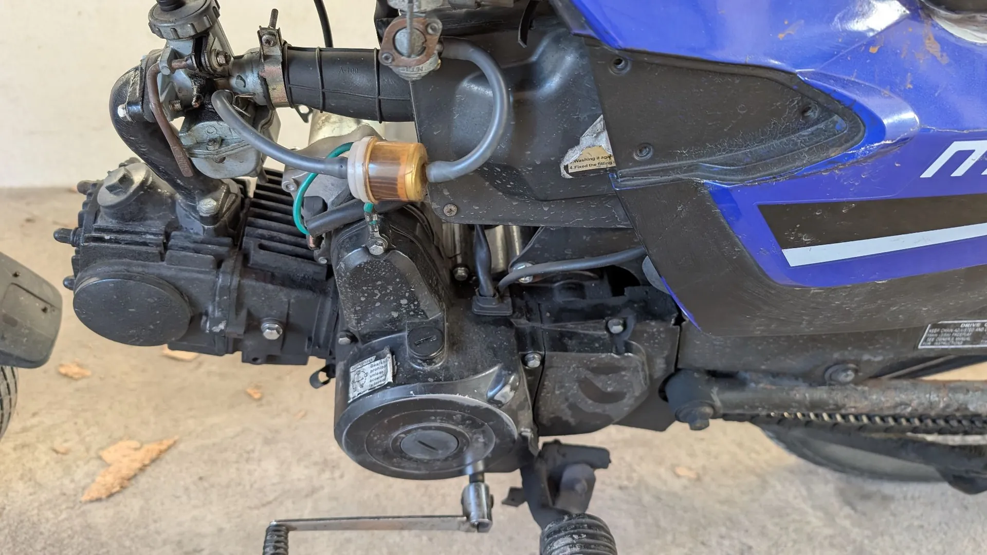 Zbliżenie na silnik i układ napędowy niebieskiego motocykla. Widoczne są czarne elementy mechaniczne, przewody, brązowy kondensator oraz fragment łańcucha napędowego po prawej stronie. Na obudowie silnika znajduje się mała, srebrna tabliczka znamionowa z tekstem.