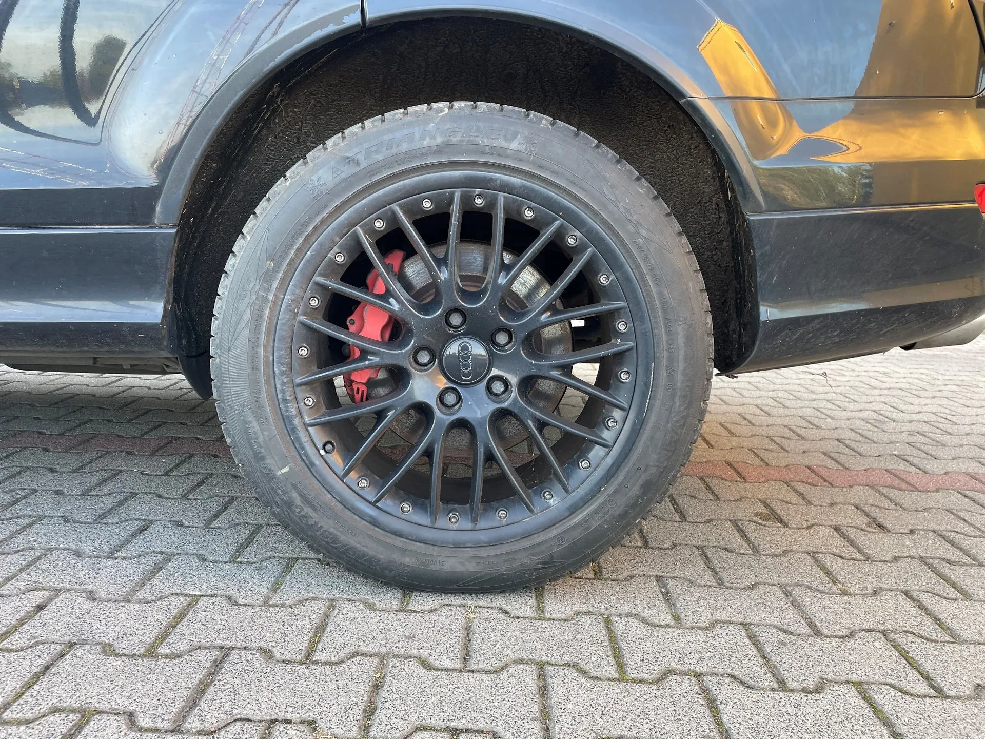Zbliżenie na czarne koło samochodu z oponą i wieloramienną czarną felgą. Przez szprychy felgi widoczny jest czerwony zacisk hamulcowy. Samochód stoi na szarej kostce brukowej.