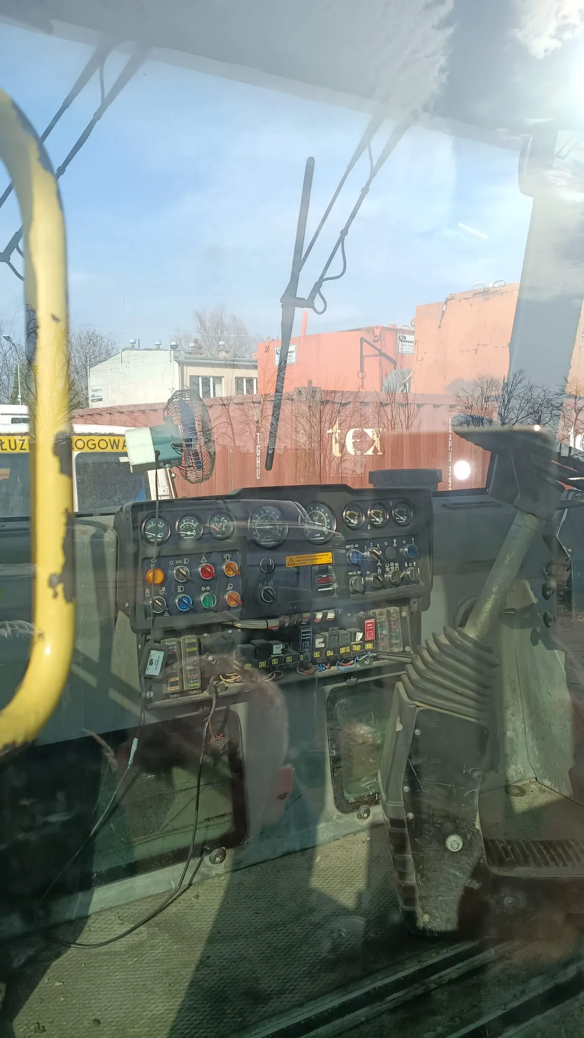 Widok z wnętrza kabiny maszyny budowlanej lub pojazdu specjalistycznego. W centrum znajduje się panel sterowania z licznymi analogowymi wskaźnikami, przełącznikami i kolorowymi przyciskami (czerwonymi, zielonymi, żółtymi). Na panelu widnieje żółta etykieta ostrzegawcza. Po prawej stronie znajduje się dźwignia sterująca z gumową osłoną przeciwkurzową. W tle, za szybą kabiny, widoczne są budynki przemysłowe w kolorze czerwonym i białym oraz fragment napisu „tex” na ścianie budynku. Po lewej stronie widoczny jest fragment żółtej ramy kabiny.