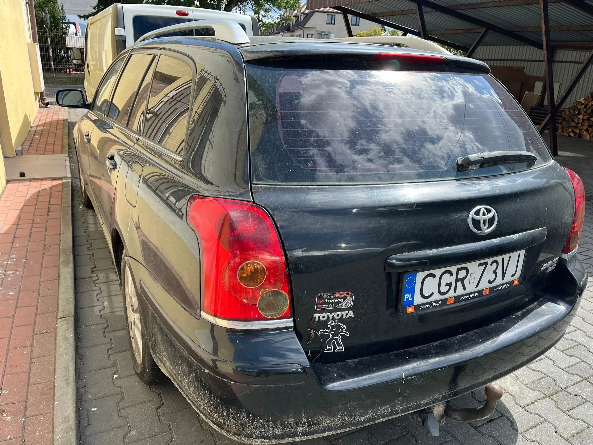 Tylny widok czarnego samochodu marki Toyota w nadwoziu kombi, zaparkowanego na kostce brukowej. Pojazd jest zakurzony, posiada polską tablicę rejestracyjną o numerze CGR 73VJ oraz kilka naklejek na klapie bagażnika, w tym logo Toyoty i grafikę postaci. W tle widoczny jest biały samochód dostawczy oraz fragmenty zabudowań.