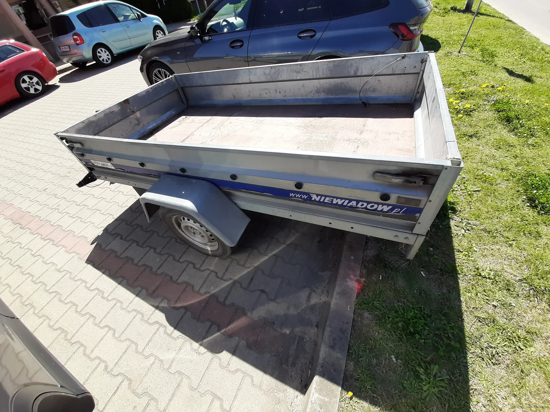 Metalowy przyczepa o prostokątnym kształcie z niebieską taśmą i napisem www.NIEWIADOW.pl na boku. Przyczepa stoi na brukowanej powierzchni obok trawnika z żółtymi kwiatami. W tle widoczne są trzy samochody: czerwony, jasnoniebieski i ciemnoszary. Cień przyczepy pada na bruk. Trawnik znajduje się po prawej stronie obrazu.