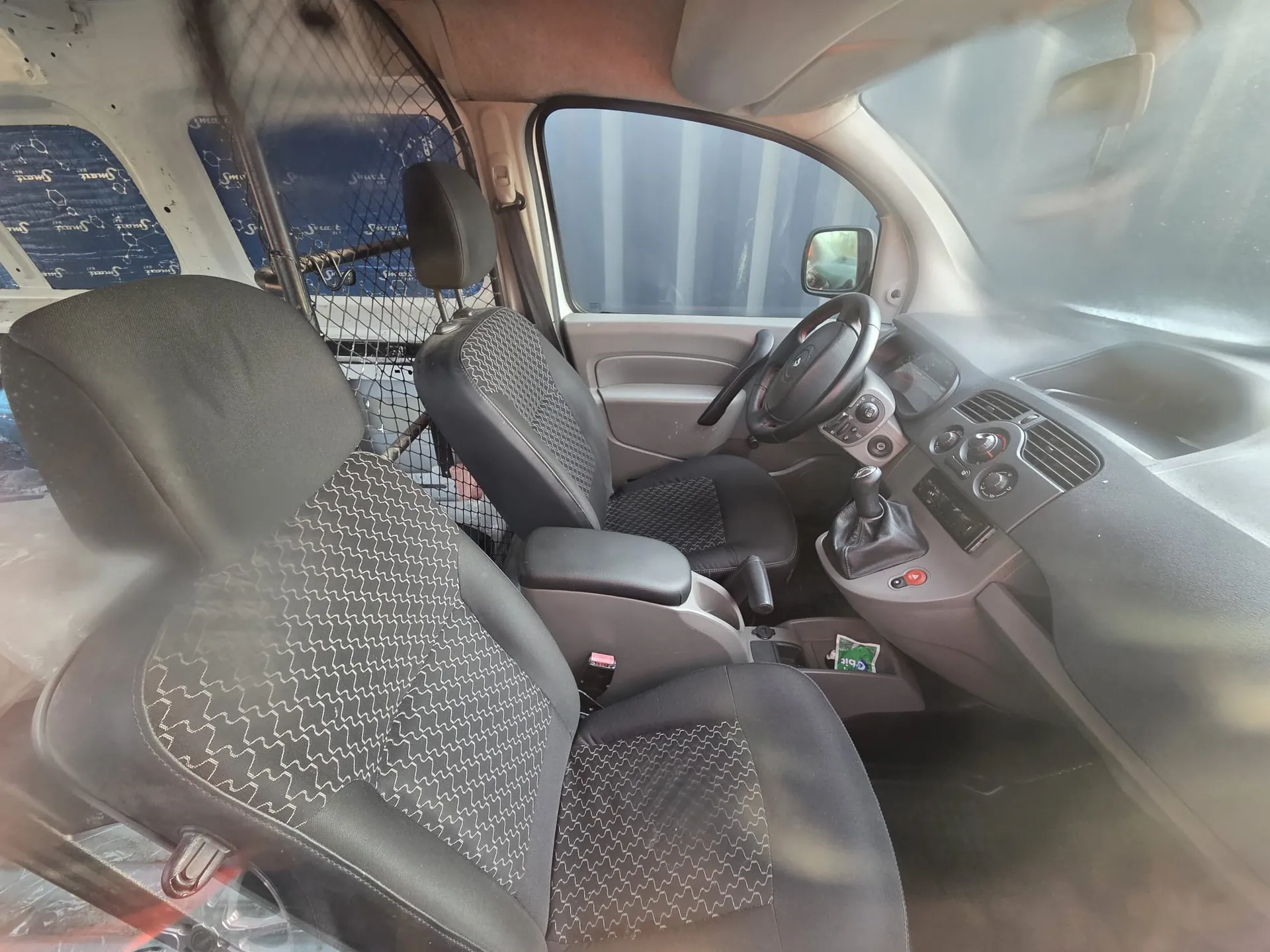 Wnętrze samochodu z widocznymi przednimi siedzeniami o czarno-białym wzorze zygzakowym. Kierownica po lewej stronie, deska rozdzielcza z przyciskami i gałkami sterującymi, dźwignia zmiany biegów. Za przednimi siedzeniami siatka oddzielająca przestrzeń załadowczą. Na bocznych szybach niebieskie panele z powtarzającym się napisem "Smart". W centralnej konsoli widoczny mały zielono-biały przedmiot.