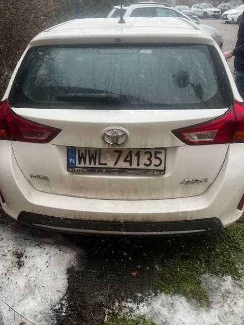 Tył białego samochodu marki Toyota Auris z tablicą rejestracyjną PL WWL 74135. Samochód jest lekko zabrudzony, widoczne są czerwone światła tylnego oświetlenia oraz logo marki. W tle inne zaparkowane pojazdy, ziemia z resztkami śniegu i mchem. Na szybie tylnej odbicia drzew.