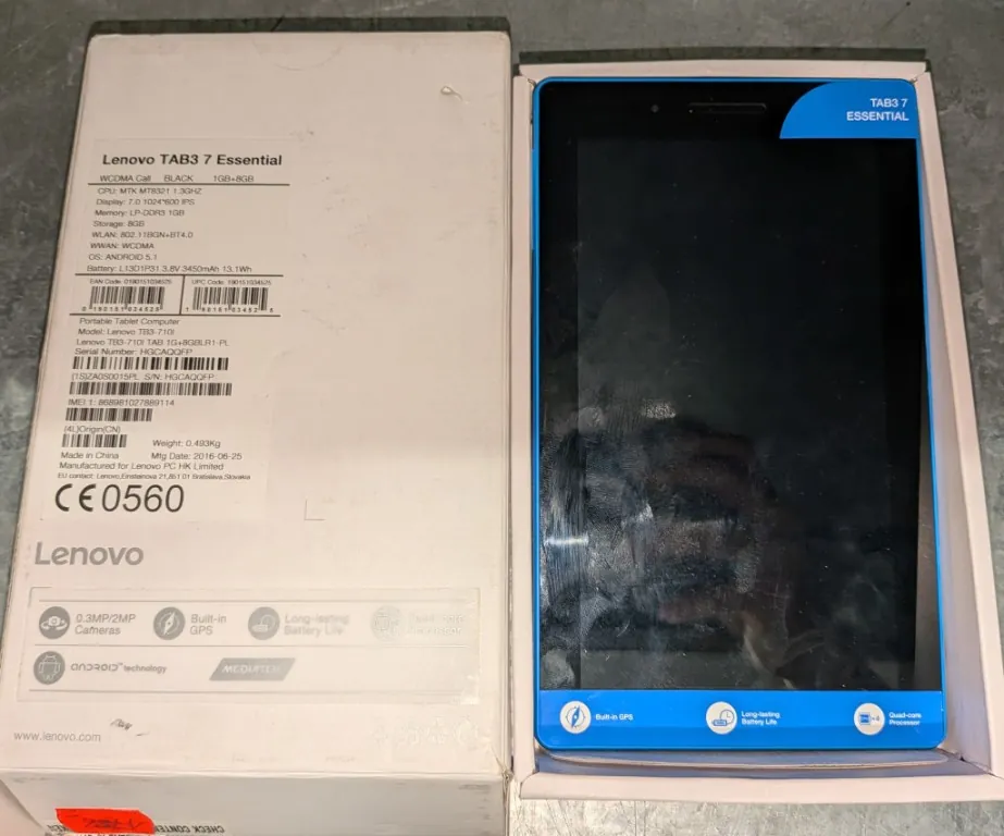 Zdjęcie przedstawiające tablet Lenovo TAB3 7 Essential w oryginalnym niebieskim opakowaniu, obok którego leży białe pudełko z etykietą techniczną. Na etykiecie widnieją napisy: 'Lenovo TAB3 7 Essential', 'WCDMA Call BLACK 1GB+8GB', specyfikacja techniczna procesora, pamięci i baterii, a także numer seryjny i oznaczenie CE 0560. Na dole pudełka znajdują się ikony funkcji: '0.3MP/2MP Cameras', 'Built-in GPS', 'Long-lasting Battery Life' oraz logo 'MediaTek'. Tablet ma czarną ramkę i ciemny ekran z widocznymi śladami użytkowania.