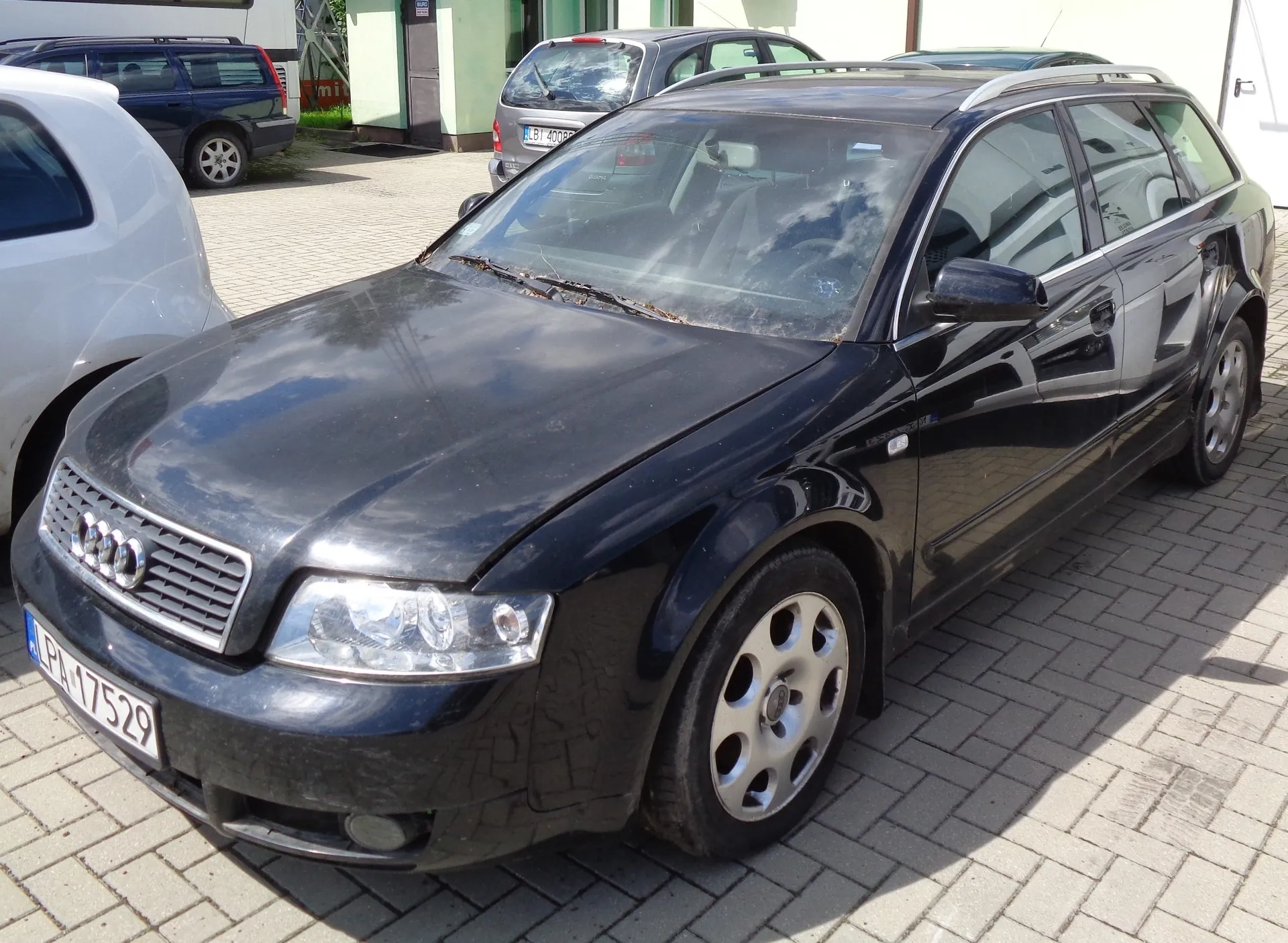 Czarny samochód marki Audi w nadwoziu kombi zaparkowany na kostce brukowej. Widoczna jest przednia część pojazdu z tablicą rejestracyjną o numerze PA 17529. W tle znajdują się inne zaparkowane samochody: biały po lewej stronie oraz srebrny i granatowy za Audi.