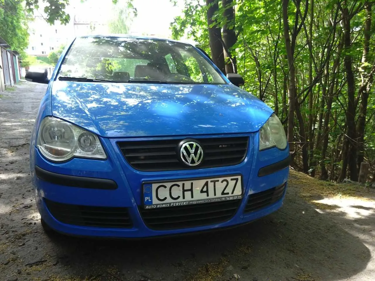 Niebieski samochód osobowy marki Volkswagen, widok z przodu. Tablica rejestracyjna z białym tłem i czarnym napisem 'CCH 4T27', po lewej stronie tablicy oznaczenie 'PL'. Poniżej tablicy napis 'AUTO KOMIS PERFECT' oraz dodatkowy tekst. Samochód zaparkowany na drodze ziemnej, w tle drzewa i budynki. Słoneczny dzień.