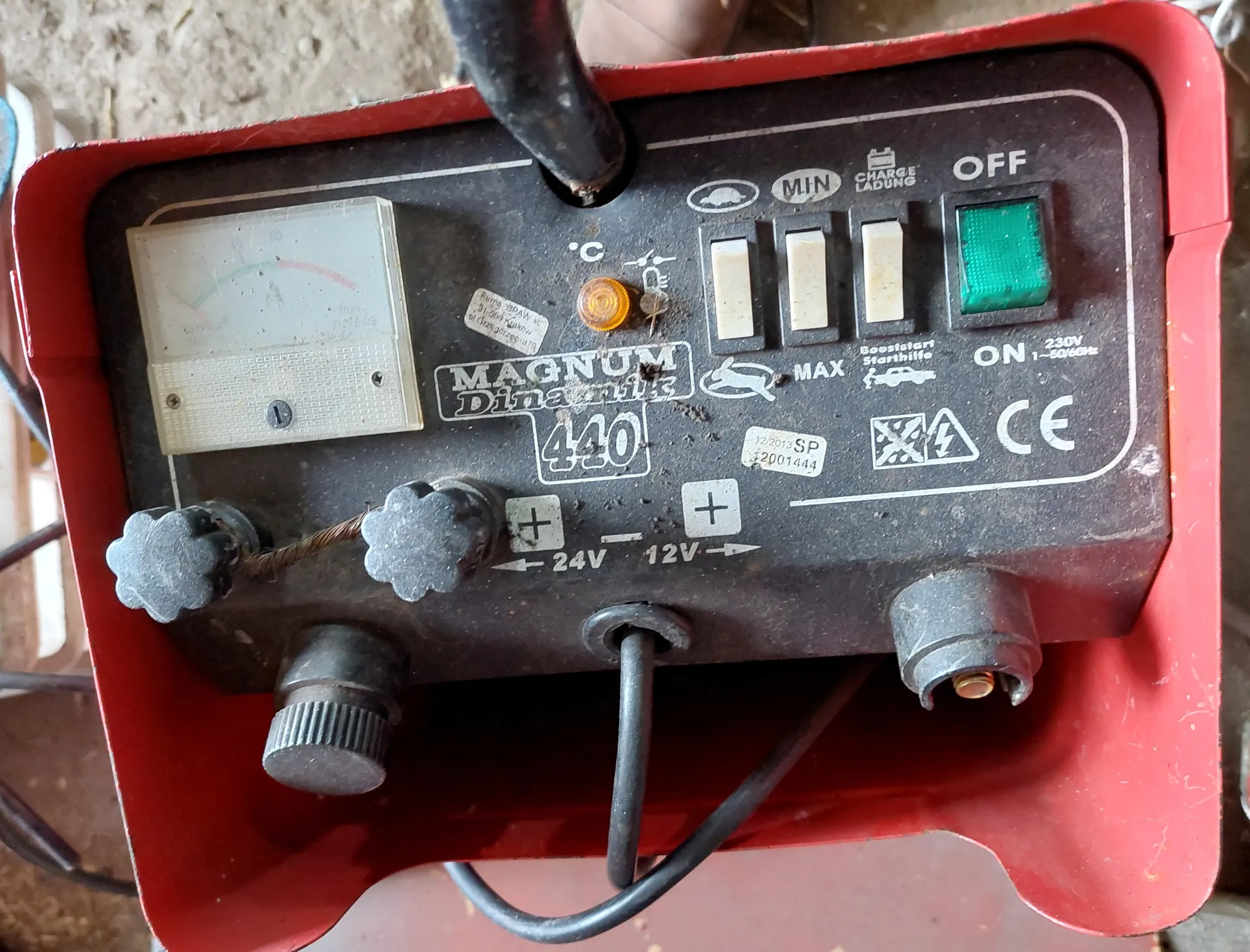 Czerwone urządzenie ładowarki akumulatorów MAGNUM Dinamik 440 z czarnym panelem sterowania. Na panelu analogowy wskaźnik, cztery przełączniki (trzy białe i jeden zielony z napisami OFF i ON), dwa czarne gałki z oznaczeniami +, etykiety: '24V', '12V', 'MIN', 'MAX', 'CHARGE LADUNG', '230V 1-50/60Hz', znak CE oraz symbole: samochód, bateria, ostrzeżenie. Widoczne przewody i elementy połączeniowe. Na panelu również napisy 'MAGNUM Dinamik 440' oraz dodatkowe naklejki z numerami.