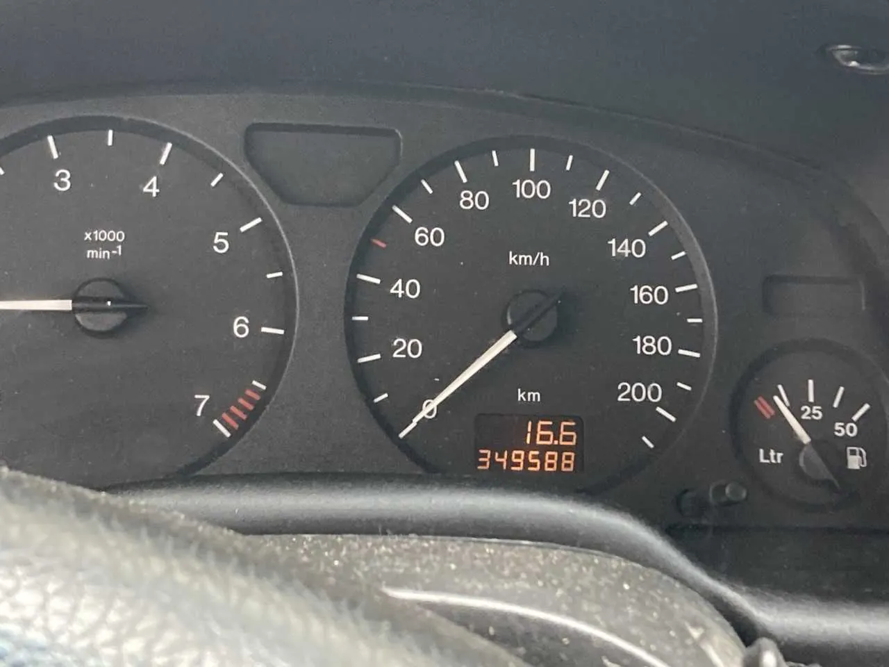 Zbliżenie na deski rozdzielcze samochodu. Po lewej stronie wskaźnik obrotomieru z zakresem od 3 do 7 (oznaczony jako x1000 min⁻¹), z czerwonymi segmentami w sekcji 6-7. W centrum wskaźnik prędkościomierza z zakresem do 200 km/h, wskazówka na 0 km/h. Poniżej cyfrowy wyświetlacz z wartościami 16.6 i 349588. Po prawej stronie wskaźnik poziomu paliwa z oznaczeniem Ltr i ikoną zbiornika, zakres do 50. Widoczny fragment kierownicy na dole obrazu. Tekst na przyrządach: km/h, km, Ltr, x1000 min⁻¹.