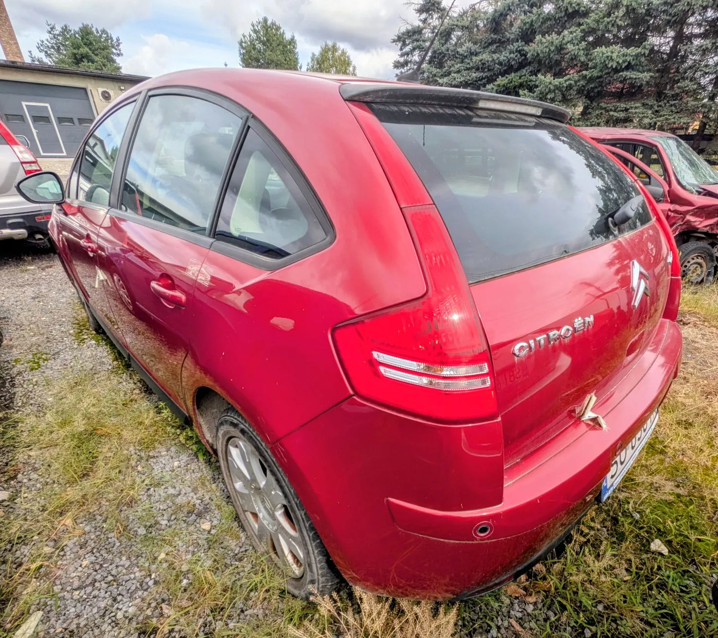 Tylny lewy trzy czwarte widok czerwonego samochodu marki Citroën zaparkowanego na żwirowym podłożu z trawą. Na klapie bagażnika widnieje napis CITROËN oraz logo marki. Samochód jest lekko zabrudzony, a w tle widać fragmenty innych pojazdów oraz drzewa.