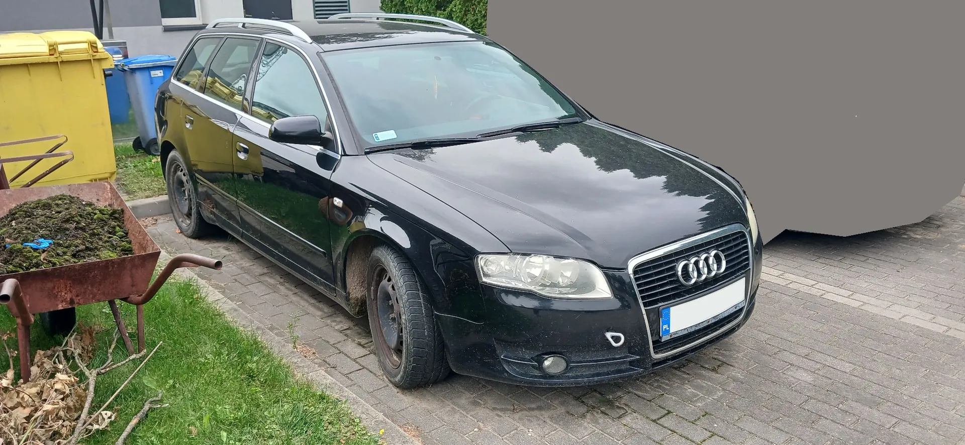 Czarny samochód marki Audi w nadwoziu kombi zaparkowany na kostce brukowej. Po lewej stronie zdjęcia widoczne są żółty i niebieski kontener na odpady oraz metalowa taczka wypełniona ziemią i roślinami. W tle znajduje się fragment budynku i zieleń.