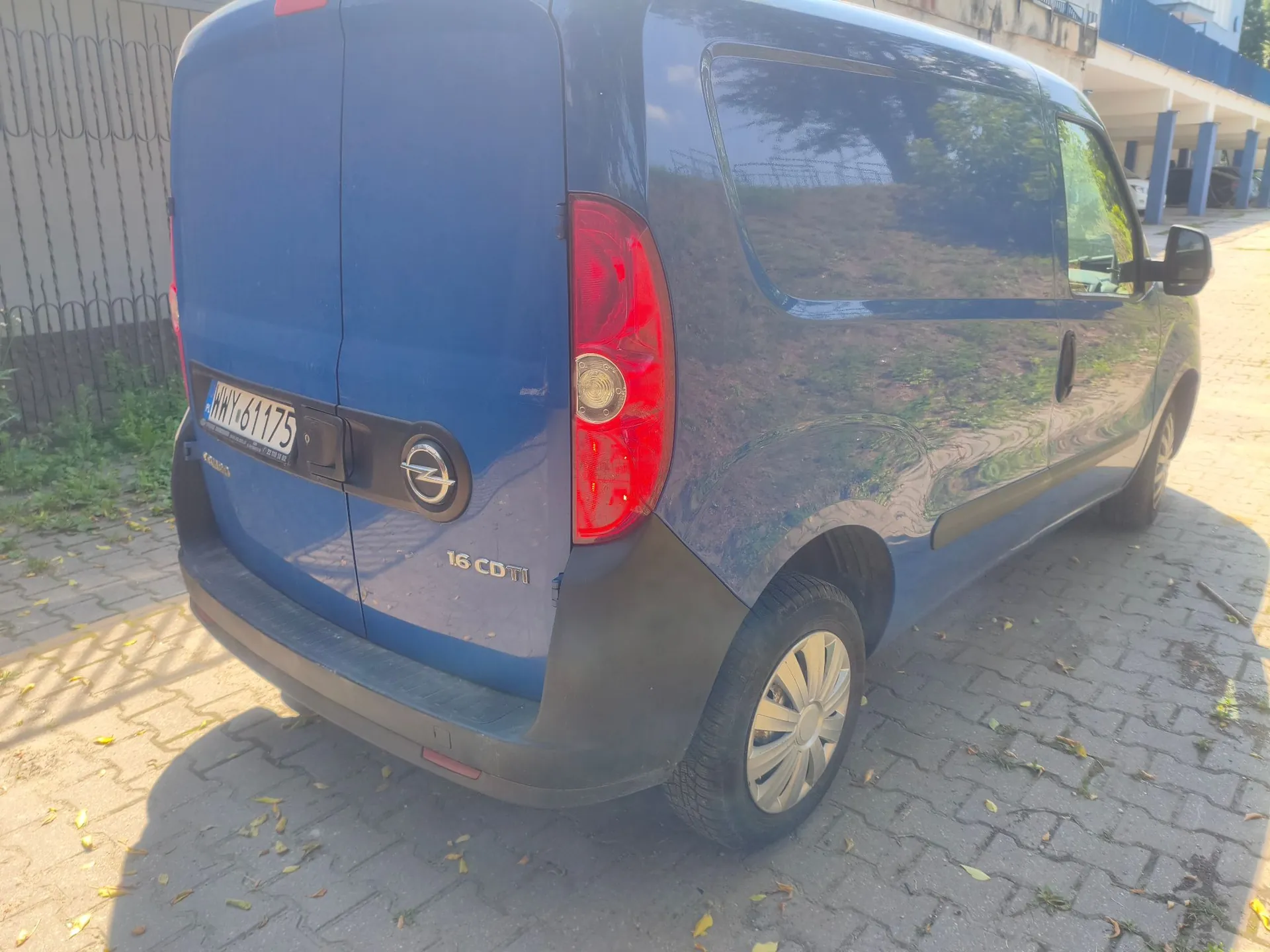 Niebieski samochód dostawczy marki Opel Combo zaparkowany na kostce brukowej. Widoczny jest tył i prawy bok pojazdu. Na klapie bagażnika znajduje się logo Opela, napis '1.6 CDTI' oraz tablica rejestracyjna o numerze 'WHY 61175'.