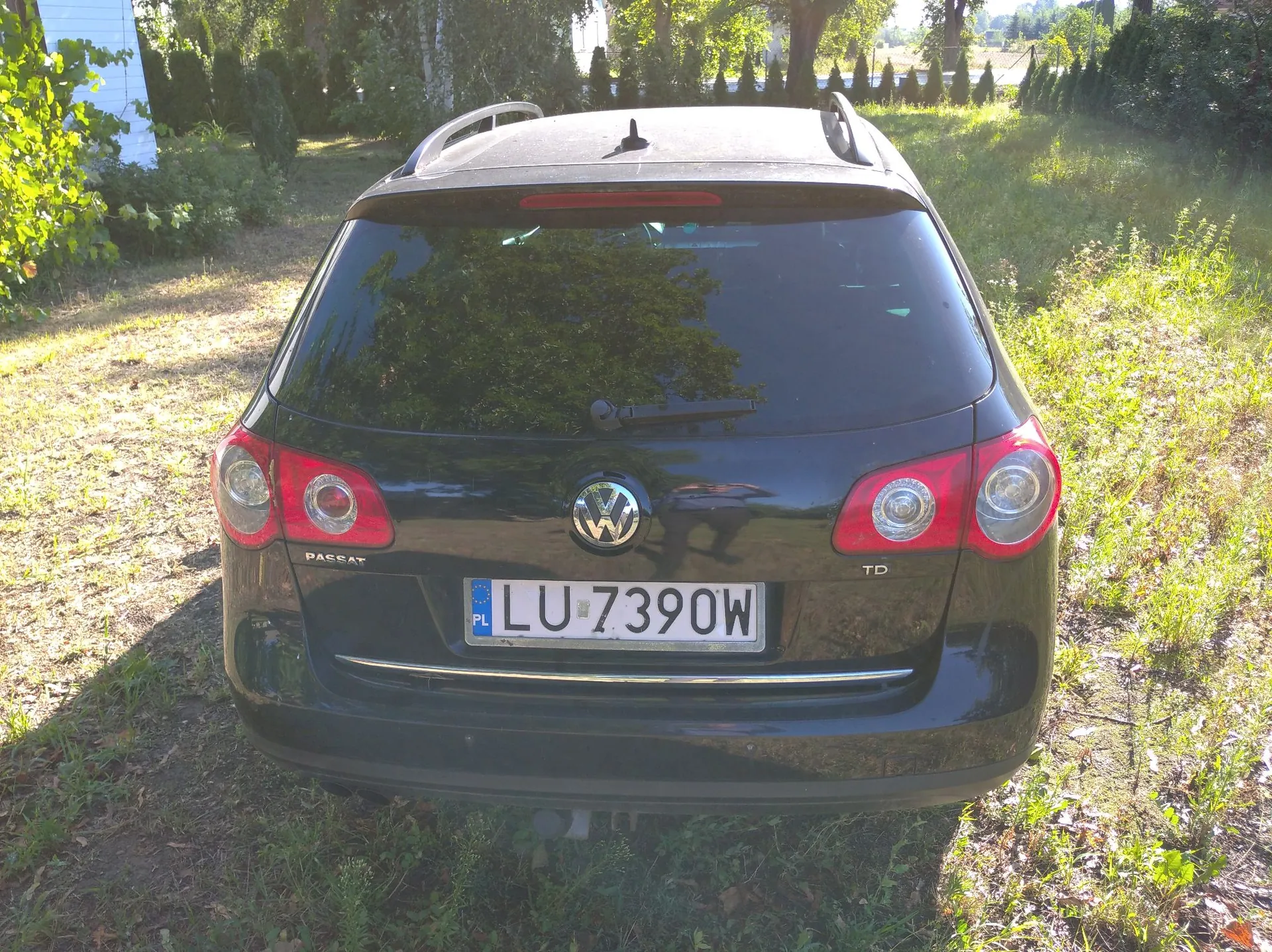 Tył czarnego samochodu marki Volkswagen Passat TD z polską tablicą rejestracyjną LU 7390W. Samochód stoi na trawiastym terenie w otoczeniu zieleni i drzew.