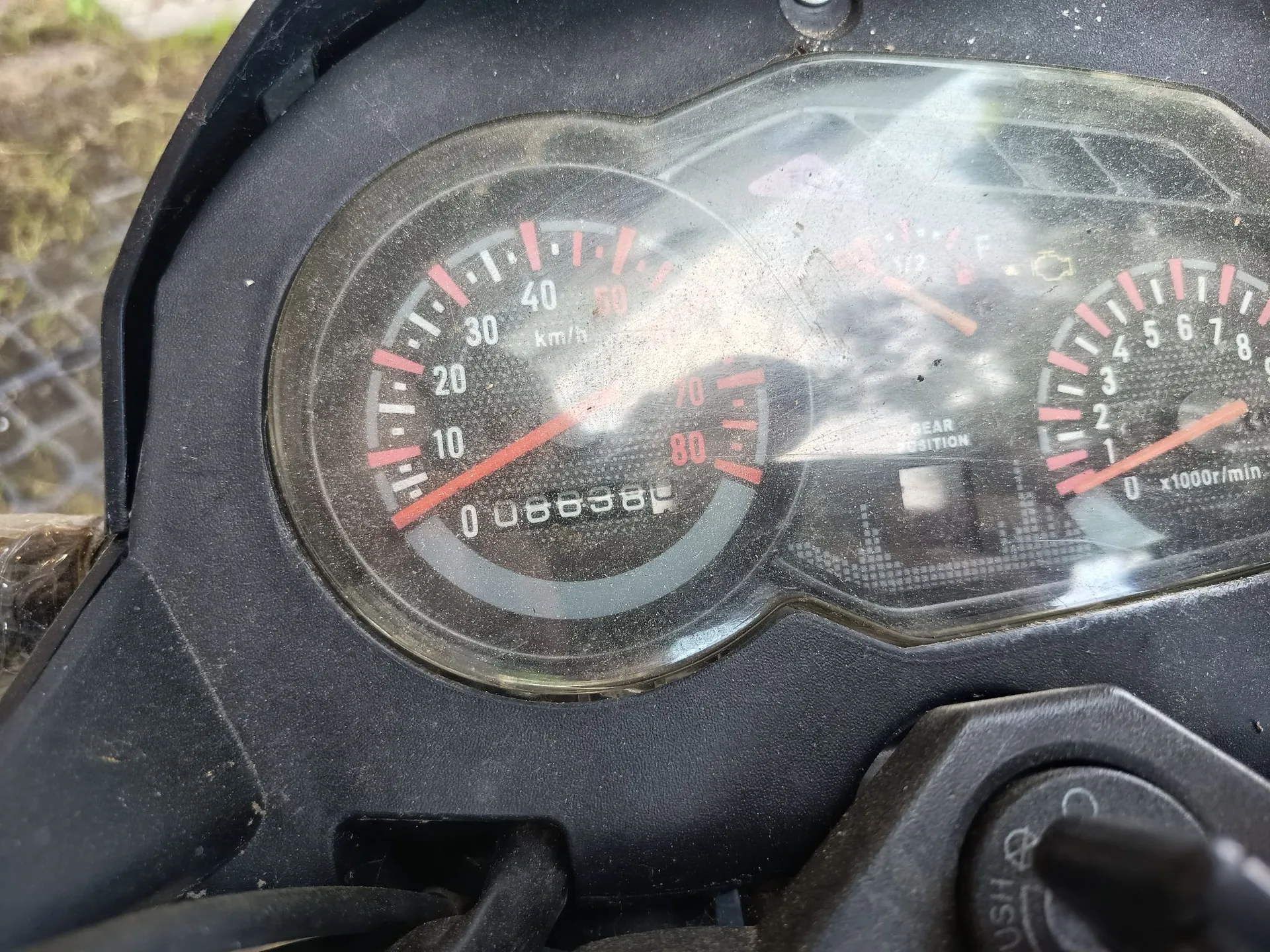 Zbliżenie na panel instrumentów motocykla. Widoczny jest analogowy prędkościomierz z podziałką do 80 km/h, wskazówka wskazuje wartość bliską 0. Poniżej znajduje się licznik kilometrów z wartością 008638. Po prawej stronie widoczny jest obrotomierz z podziałką od 0 do 8 (x1000r/min) oraz wskaźnik poziomu paliwa. Całość znajduje się pod lekko zabrudzoną, przezroczystą szybką w czarnej obudowie.