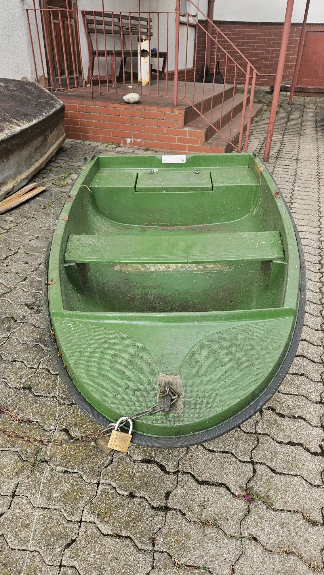 Zielona, plastikowa łódka o owalnym kształcie, ustawiona na kostce brukowej przed budynkiem. Łódka posiada wewnątrz dwie poziome półki/siedziska. Przednia część łodzi jest zabezpieczona łańcuchem z kłódką, który jest przymocowany do metalowego uchwytu. W tle widoczne są schody z ceglanymi stopniami, metalowa barierka oraz fragment białej ściany budynku.
