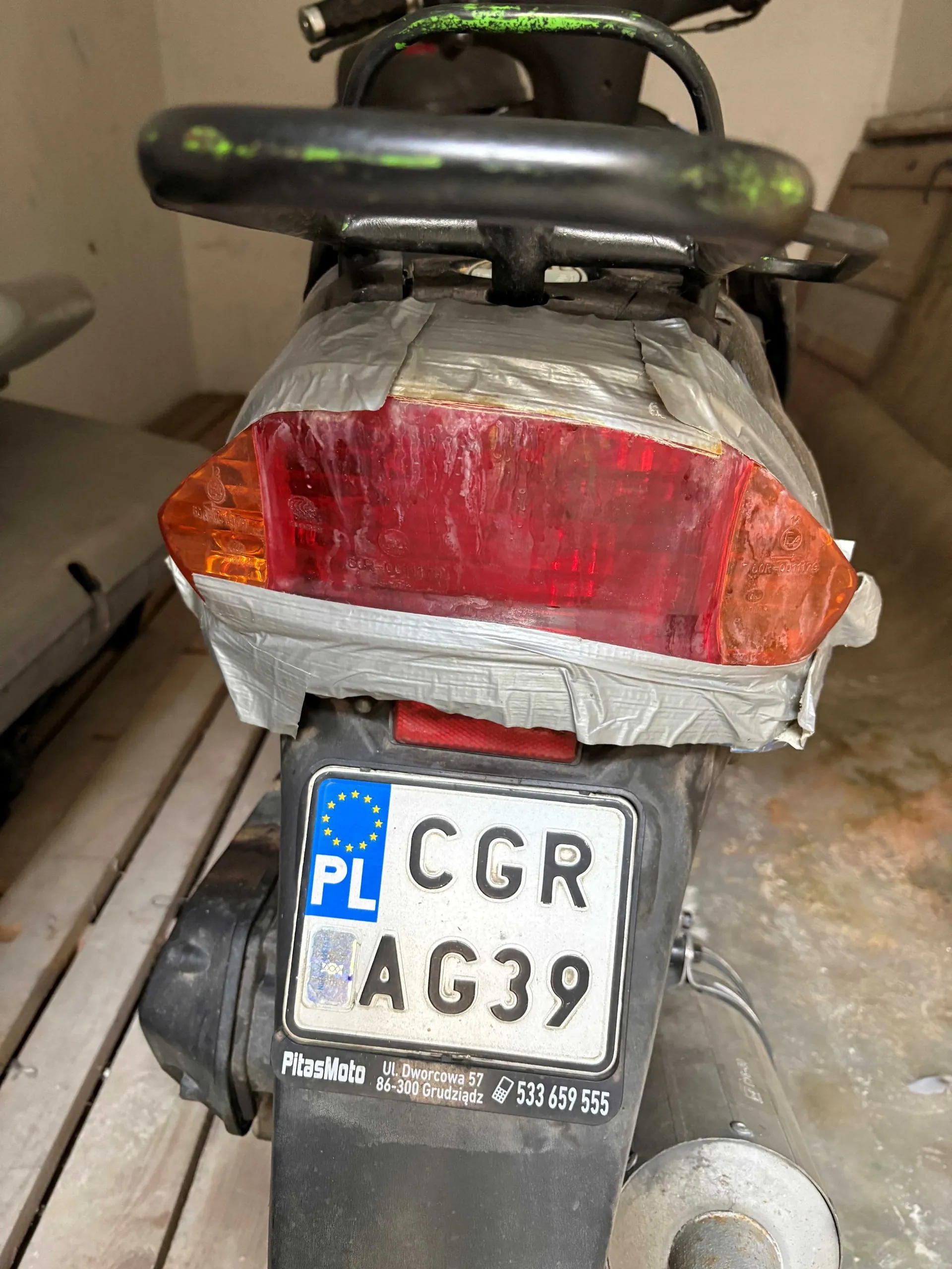 Widok z tyłu na motocykl lub skuter. Tylna lampa jest owinięta szeroką, szarą taśmą klejącą. Poniżej znajduje się polska tablica rejestracyjna o numerze CGR AG39. Pod tablicą widnieje naklejka z napisem 'PitasMoto UL. Qworcowa 57 84-300 Grudziądz' oraz numerem telefonu '533 659 555'.