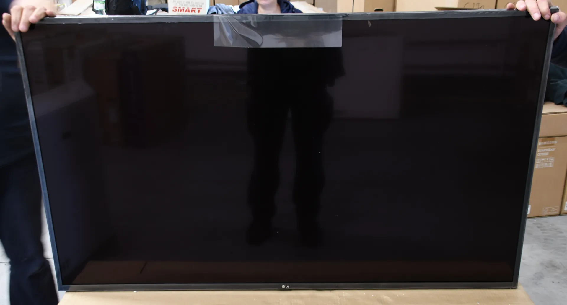 Osoba trzymająca duży, wyłączony telewizor z czarnym ekranem. Na górnej krawędzi ekranu znajduje się kawałek przezroczystej taśmy klejącej. Na dolnej ramce widoczne jest logo LG. W tle znajdują się kartonowe opakowania.