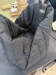 Ciemnoszara pikowana kurtka leżąca na ciemnej powierzchni. Widoczna zielona etykietka. W tle fragment pomieszczenia z drewnianymi patykami i jasną podłogą.