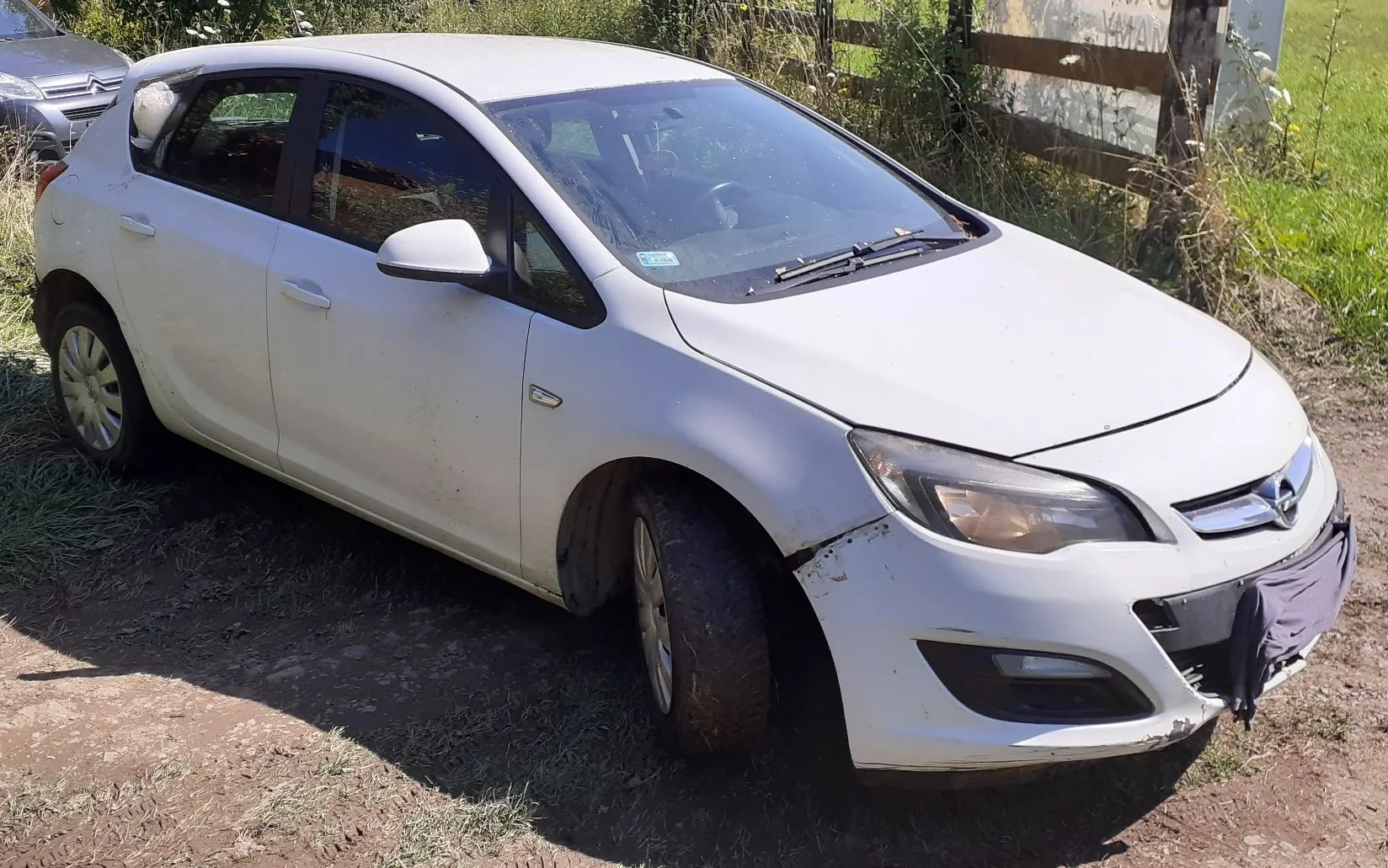 Biały samochód osobowy marki Opel Astra w nadwoziu hatchback, zaparkowany na nieutwardzonym, trawiastym terenie. Pojazd jest zabrudzony, a na przednim prawym zderzaku widoczne są uszkodzenia i wgniecenia. Przedni pas samochodu jest częściowo zakryty szarą tkaniną.