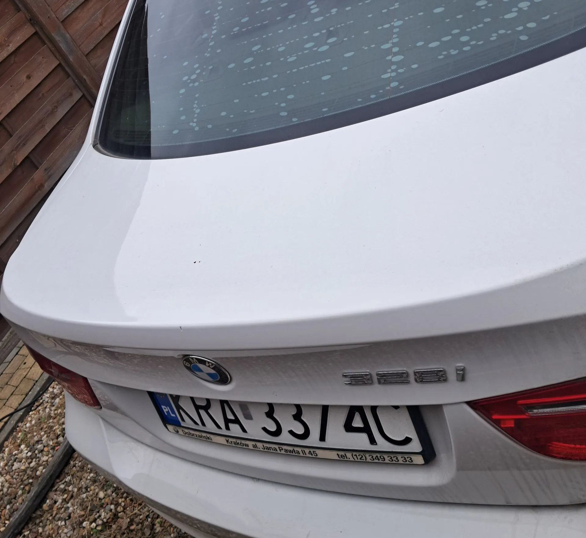Tylna część białego samochodu marki BMW, model 320i. Widoczna jest tylna szyba z kropkami odgrzewacza, logo BMW oraz tablica rejestracyjna o numerze KRA 3374C. Poniżej numeru rejestracyjnego znajduje się napis: 'Dobrzalinski Kraków al. Jana Pawła II 45 tel. (12) 349 33 33'. Samochód zaparkowany jest obok drewnianego ogrodzenia.