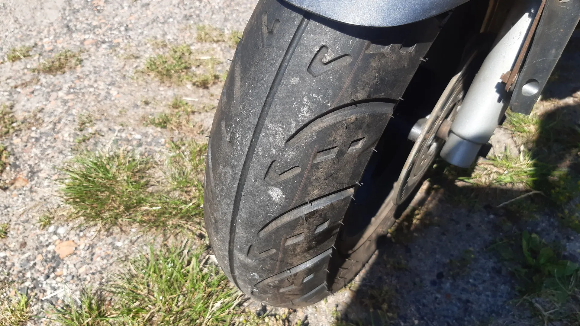 Zbliżenie na przednią oponę motocykla lub skutera, ustawioną na szarej, żwirowej nawierzchni z kępkami zielonej trawy. Opona jest czarna, z widocznym bieżnikiem i śladami zużycia oraz zabrudzeń. W tle widoczny jest fragment srebrnego zawieszenia i błotnika.